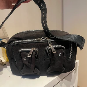 Svart axelväska - Nunoo - Cool svart axelväska med bred axelrem i skinn och flera silvriga dragkedjor. Väskan har två praktiska fickor framtill med detaljer i metall och robust tygmaterial. Perfekt för dig som gillar streetstyle och vill ha plats för allt viktigt.
