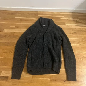 Mörkgrå kofta från Dressmann - Mysig mörkgrå kofta från Dressmann med sjalkrage och knäppning framtill. Koftan är ribbstickad och har två praktiska fickor framtill. Perfekt för lager-på-lager och chill dagar. Passar dig som gillar klassisk och enkel stil.