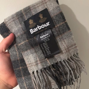 Rutig grå halsduk från Barbour - Snygg rutig halsduk från Barbour i olika nyanser av grått och beige med inslag av brunt. Gjord i 100% ull och har klassiska fransar i kanterna. Perfekt accessoar för att höja din stil under kyliga dagar.