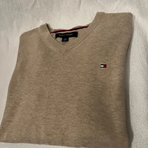 Beige v-ringad tröja Tommy Hilfiger - Snygg beige v-ringad tröja från Tommy Hilfiger i mjuk bomull. Klassisk design med diskret logga broderad på bröstet och ribbade muddar vid halsen. Perfekt för dig som gillar stilrena och tidlösa plagg.