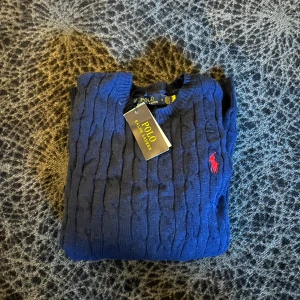 Marinblå stickad tröja Polo Ralph Lauren - Snygg marinblå stickad tröja från Polo Ralph Lauren i storlek S. Tröjan har klassiskt kabelstickat mönster och en röd broderad logga på bröstet. Tillverkad i 100% bomull med rund hals och ribbade muddar. Perfekt för dig som gillar stilrena och tidlösa plagg.