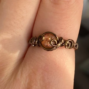 Handgjord ring med glittrig sten - Unik handgjord ring i bronstonad metall med en glittrig orange sten i mitten. Ringen har snygga snirkliga detaljer runt hela bandet som ger den en bohemisk och cool vibe. Perfekt för dig som gillar statement-smycken och vill sticka ut lite extra.