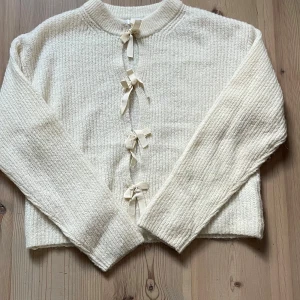 Vit stickad kofta med rosetter - Superfin vit stickad kofta med flera söta rosetter framtill. Koftan har långärmade ärmar och en mjuk, ribbad struktur som gör den extra mysig. Perfekt för dig som gillar gulliga detaljer och vill ha en cozy look.