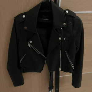 Svart croppad skinnjacka med bälte - Snygg svart croppad skinnjacka från pull & bear med silvriga dragkedjor, knappar och bälte i midjan. Använd 1 gång. 