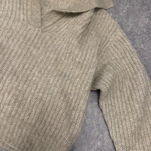 Beige stickad tröja från Gina Tricot - Säljer en beige stickad tröja från Gina Tricot med krage och v-ringad hals. Tröjan har långärmade och ribbad struktur som ger en chill och snygg vibe. Perfekt att slänga på sig när det är lite kyligt ute.