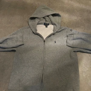 Grå hoodie från Polo Ralph Lauren XL - Grå hoodie från Polo Ralph Lauren i storlek XL med klassisk logga broderad på bröstet. Tröjan har huva, dragkedja framtill och två fickor. Tillverkad i mjuk bomullsmix, perfekt för chill dagar. Snygg och enkel stil som funkar året runt.