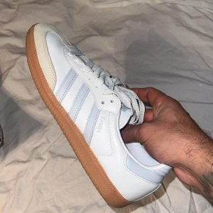 Adidas Samba sneakers vit/blå - Säljer ett par klassiska Adidas Samba sneakers i vitt och ljusblått med gummisula. Skorna har de ikoniska tre ränderna på sidan, snörning och rund tå. Ovandelen är i skinn och mocka, perfekt för dig som gillar retrostil och streetwear.