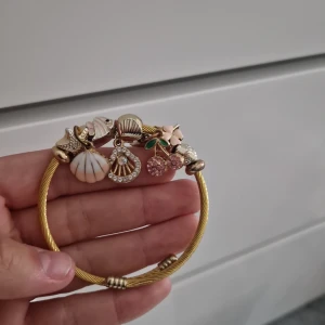 Guldigt armband med berlocker - Charmigt guldigt armband med flera berlocker, bland annat snäckor, körsbär, blomma och fisk. Berlockerna har detaljer i vitt, rosa, grönt och glittrande stenar. Perfekt accessoar för dig som gillar unika smycken med somrig känsla. Köpt i spanien