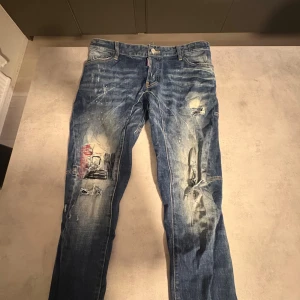Dsquared2 blå jeans med slitningar - Snygga blå jeans från Dsquared2 med coola slitningar, färgstänk och patchade detaljer. Jeansen har klassisk femficksdesign, tydliga kontrastsömmar och en unik look med tryck och slitna partier på framsidan. Perfekt för dig som gillar streetstyle.