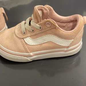 Ljusrosa Vans sneakers med vit sula - Säljer ett par snygga Vans sneakers i ljusrosa canvas med klassisk vit våg på sidan och vit gummisula. Skorna har snörning och rund tå, samt den typiska bruna mönstrade yttersulan. Perfekt för dig som gillar streetstyle och vill ha en fräsch look.