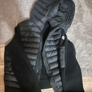 Svart Moncler pufferjacka med huva - Säljer en svart pufferjacka från Moncler med stickade ärmar och quiltade partier. Jackan har en stor huva, dragkedja framtill och Moncler-logga på ärmen. Perfekt för kyliga dagar och riktigt snygg till streetwear-stilen.