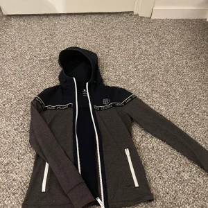 Grå och marinblå jacka Maya Delorez - Snygg jacka från Maya Delorez i grått och marinblått med huva och vita dragkedjor. Jackan har logga på bröstet och band med texten 'Maya Delorez' över axlarna. Perfekt för dig som gillar sportig stil och vill sticka ut lite extra. Priset kan diskuteras är ganske snäll!