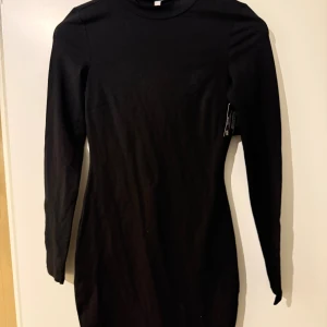 Svart långärmad klänning från H&M - En svart, figurnära klänning från H&M med lång ärm och enkel design. Klänningen har en hög halsringning och är tillverkad i ett mjukt bomullsmaterial. Perfekt basplagg att styla på många olika sätt.