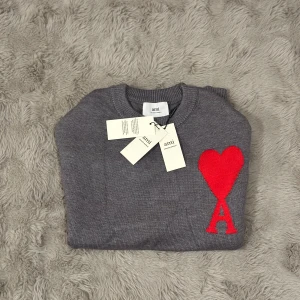 Grå stickad tröja med rött hjärta från Ami - Supersnygg grå stickad tröja från Ami Alexandre Mattiussi med ett stort rött hjärta och bokstaven A broderat på bröstet. Tröjan har rund hals och ribbade muddar för en clean look. Perfekt för dig som gillar stilrena detaljer och vill sticka ut.