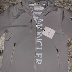 Moncler Winbreaker  - Superfin Moncler windbreaker i ljusgrått med luva, dragkedjor och stort Moncler-tryck framtill. Aldrig använd – skick 11/10! Perfekt för regn och blåst, vatten rinner direkt av tack vare det vattenavvisande materialet. Stilren, exklusiv och bekväm. Qr verifierng finns för fler bilder så är det bara att hoojta till