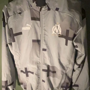 Grå vindjacka Puma x Marseille - Snygg grå vindjacka från Puma i samarbete med Marseille. Jackan har ett grafiskt mönster med mörka kors, dragkedja framtill och huva. Marseille-logga och Puma-tryck på bröstet. Perfekt för dig som gillar sportig streetstyle.