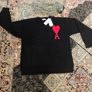 Ami Paris tjock tröja - Svart stickad tröja från Ami Paris med röd hjärtapplikation och bokstaven A på bröstet. Tröjan har rund hals, ribbade muddar och lång ärm. Perfekt för dig som gillar stilrena plagg med en unik detalj. Snygg att matcha med jeans eller kjol.