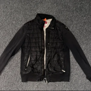 Svart quiltad jacka Parajumpers - Svart quiltad jacka från Parajumpers med cool badge på ärmen och dragkedjor i silver. Jackan har långärmade svarta ärmar i mjukt material och en quiltad front i syntet. Insidan är ljus och har märkets logga. Ny köpt för 5500 kr och är i använt skick har ett litet fel därav låg pris priset kan diskuteras