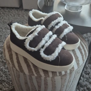 Bruna sneakers med fuskpäls från Duffy - Mysiga bruna sneakers från Duffy med vit fuskpäls längs kanterna och insidan. Skorna har breda kardborreband och en tjock vit sula. Utsidan är i mockaliknande material och de har en rund tå. Perfekta för dig som vill ha både stil och komfort.