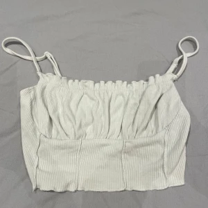 Vit ribbad linnetopp med smala axelband - Säljer en vit ribbad linnetopp med smala axelband och lätt rynkad överdel. Toppen är croppad och har en mjuk, stretchig känsla som gör den skön att bära. Perfekt för varma dagar och enkel att matcha med jeans eller kjol.