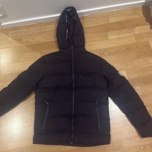 Svart Moncler pufferjacka med huva - Svart pufferjacka från Moncler med huva och logotyp på ärmen. Jackan har dragkedja framtill, två fickor med dragkedja och Moncler-tryck på insidan av huvan. Perfekt för kalla dagar och har en clean, modern look. Hål i fickorna 
