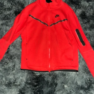 Röd Nike tech med huva  - Nike tech i rött med svarta detaljer och dragkedja framtill. Koftan har huva, lång ärm och en cool svart detalj på ena ärmen. Reflekterande band över bröstet och fickor på sidorna. Perfekt för dig som gillar sportig stil.