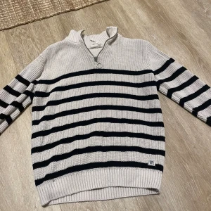 Randig stickad tröja med half zip - Snygg stickad tröja från SPRAY i ljusbeige med svarta ränder. Tröjan har långärmad design, ribbade muddar och en cool half zip-dragkedja vid halsen. Perfekt för dig som gillar klassisk stil med en modern twist.