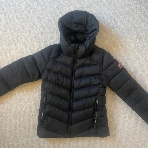 Svart pufferjacka från Superdry - Svart pufferjacka från Superdry. Jackan har huva och broderad logga på ärmen. Jackan har två dragkedjefickor fram och snygg passform till midjan. Perfekt för kalla dagar då jackan verkligen håller värmen. Skick 9/10. Storleken är liten så mer en 36:a än 38. Skicka dm vid frågor. 
