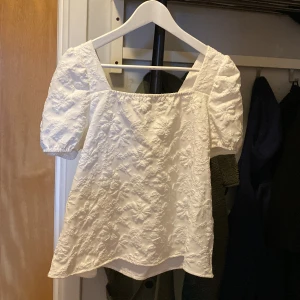 Vit blommig blus med puffärm och rosett - Supersöt vit blus med broderade blommor och puffiga korta ärmar. Blusen har en fyrkantig halsringning och en svart rosett i ryggen som ger en extra detalj. Perfekt för dig som gillar romantisk och trendig stil. Storlek S-M