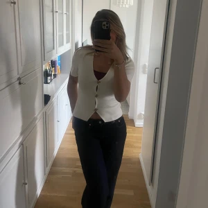 Vit kortärmad blus med knappar - Säljer en vit blus med korta ärmar och snygg v-ringning. Blusen har fyra dekorativa knappar framtill och är lätt croppad i modellen. Perfekt för dig som gillar stilrena och enkla toppar med en modern touch.