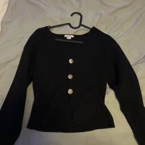 Svart v-ringad topp med knappar - Snygg svart topp från gina med v-ringning och tre stora dekorativa knappar framtill. Modellen är långärmad och har en croppad passform som ger en modern siluett. Perfekt för dig som gillar stilrena och enkla plagg med en twist. Använd endast en gång. 