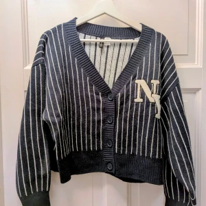 Randig marinblå kofta med NY-emblem - Snygg croppad marinblå kofta från H&M med vita ränder och stort NY-emblem på bröstet. Koftan har ribbad v-ringad krage, fyra knappar framtill och långa ärmar. Materialet är stickat och känns mjukt mot huden. Perfekt för dig som gillar college-vibe och streetstyle.