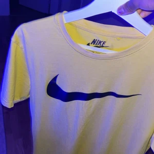 Gul Nike t-shirt med stor logga - Gul t-shirt från Nike med stor svart swoosh-logga på bröstet och en mindre logga i nacken. Klassisk rund hals och korta ärmar. Perfekt för dig som gillar sportig och clean stil.