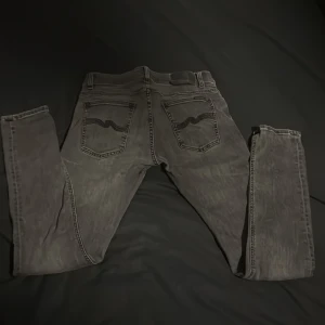 Svarta jeans från Nudie Jeans, 32/32 - Säljer ett par svarta jeans från Nudie Jeans i storlek 32/32. Jeansen har klassisk femficksdesign, svarta sömmar och Nudie-loggan på bakfickorna. Modellen är rak med slim passform och tillverkad i mjukt denimtyg.