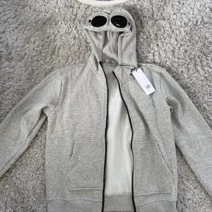 Grå hoodie med goggles från C.P. Company - Säljer en grå hoodie från C.P. Company med ikoniska goggles i huvan. Tröjan har dragkedja, två fickor fram och broderad logga på bröstet. Perfekt för dig som vill sticka ut med streetwear-vibe. Materialet är mjuk bomull och passformen är normal. Riktigt bra skick bara att det är en knapp som är sönder men inga stora problem 