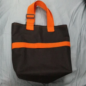 Svart shoppingväska med orange detaljer - Cool svart shoppingväska med starka orange handtag och ett orange band runt väskan. Väskan är tillverkad i slitstark polyester och har en enkel, boxig form som rymmer mycket. Perfekt för att bära med sig allt man behöver under dagen. Väskan är ett återbruk av en gammal stativväska så den kan ha lite skavanker. Den är 33 cm bred och ca 30 cm hög. Ca 10 cm i botten. 