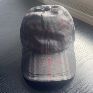 Rutig keps från Burberry i bomull - Säljer en klassisk rutig keps från Burberry i svart, grått och rött. Kepsen har den ikoniska Burberry-rutiga designen och är tillverkad i 100% bomull. Perfekt accessoar för en stilren och trendig look.