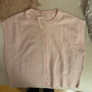 Ljusrosa stickad tröja  från Nelly - Supersöt ljusrosa stickad tröja  från Nelly med rund halsringning och ribbade kanter. Tröjan  har knappar framtill i matchande färg och är ärmlös med en loose fit. 