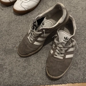 Adidas Samba grå/vita sneakers - Adidas Samba sneakers i grå mocka med vita detaljer och klassiska tre ränder på sidorna. Skorna har snörning, vit plös med svart Adidas-logga och en platt sula med hexagonmönster undertill. Perfekta för en sportig och avslappnad stil. 