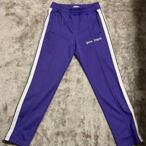 Palm Angels lila trackpants M - Snygga lila trackpants från Palm Angels med vita sidoränder och logga på låret. Byxorna har elastisk midja, raka ben och är gjorda i 100% polyester för en sportig vibe. Perfekta för dig som gillar streetwear och vill sticka ut med färg.
