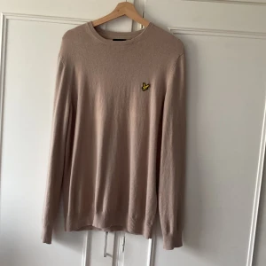 Beige stickad tröja från Lyle & Scott - ANVÄND 1 gång! Snygg beige stickad tröja från Lyle & Scott med rund hals och klassisk gul logga på bröstet. Tröjan har långa ärmar och ribbade muddar vid ärmslut och nederkant. Perfekt tröja under hösten och vintern.