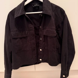 Svart overshirt från Monki, stl S - Svart overshirt från Monki i bomull med klassisk krage, två stora bröstfickor och silvriga knappar framtill. Jackan har en croppad passform och långa ärmar med knapp vid ärmslut. Perfekt för lager-på-lager och enkel att styla.