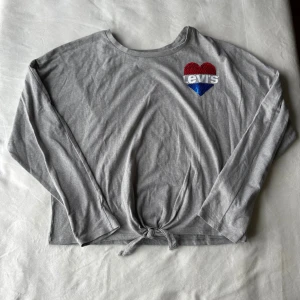 Grå Levi's tröja - Grå långärmad tröja från Levi’s med ett paljetthjärta framtill. Storlek: 165cm (16A). Fraktar inom 24h!