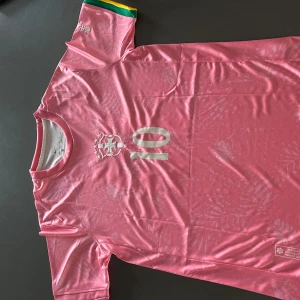 Rosa Neymar Jr fotbollströja - Snygg rosa fotbollströja från brasilien med Neymar Jr och nummer 10 på ryggen. Tröjan har korta ärmar, broderad logga och subtilt mönster i tyget. Detaljer i gult och grönt på ena ärmen. 10/10 kvalitet. Ber om ursäkt för de dåliga bilderna.