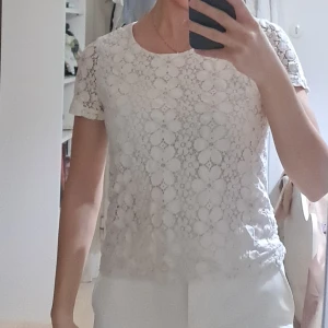 Vit spetsblus från Vero Moda XS - Supersöt vit blus från Vero Moda i storlek XS. Blusen har korta ärmar och är täckt av ett blommigt spetsmönster som ger en romantisk vibe. Rund halsringning och fodrad framtill för extra komfort. Perfekt att styla med jeans eller kjol!