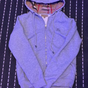 Grå hoodie från Burberry med dragkedja - Säljer en grå hoodie från Burberry med klassiskt rutigt foder i huvan och broderad logga på bröstet. Tröjan har snörning i huvan, ribbade muddar och dragkedja framtill. Perfekt för dig som gillar stilrena och bekväma plagg.