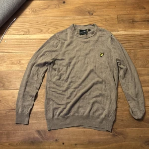 Beige stickad tröja från Lyle & Scott - Säljer en beige stickad tröja från Lyle & Scott med klassisk rund hals och den ikoniska gula fågel-loggan broderad på bröstet. Tröjan har långa ärmar och ribbade muddar för en snygg passform. Perfekt för lager-på-lager under kyliga dagar. Storlek S, fint skick