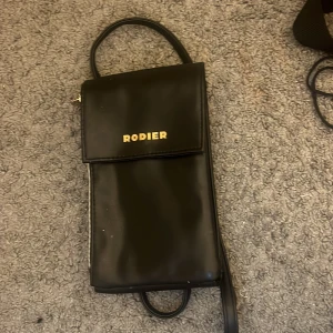 Svart axelväska från Rodier i skinn - Snygg svart axelväska från Rodier i slätt skinn med guldiga detaljer och dragkedja på sidan. Väskan har ett stilrent, avlångt format och lock med magnetknäppning. Perfekt för mobilen och småsaker när du är på språng.