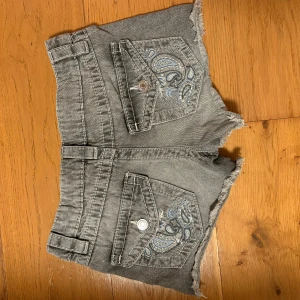 Grå denimshorts med broderade fickor - Snygga grå jeansshorts med slitna kanter och broderade detaljer i blått och silver på bakfickorna. Modellen är låg midjade/mid waist med knapp och dragkedja framtill, samt bälteshällor. Perfekta för sommarens chill dagar 💕