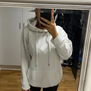 Vit hoodie med huva och snörning - Säljer en vit hoodie med huva och snörning framtill. Tröjan har en stor magficka och är långärmad med ribbade muddar vid ärmslut och nederkant. Perfekt för chill dagar och enkel att matcha med jeans eller leggings.